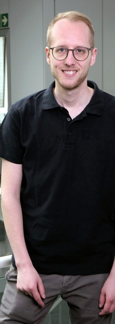 Bild vom Gründer Hendrik Brettschneider Lächelnder Mann mit Brille in einem hellblauen Hemd, steht in modernem Büro.
